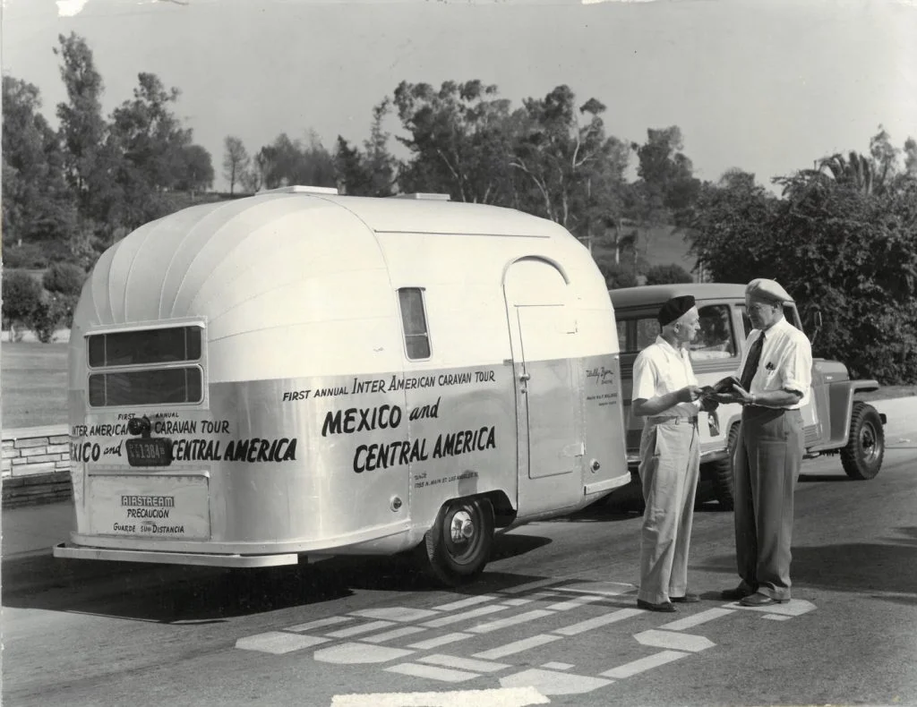 Black and white photo of two men talking beside a vintage travel trailer hitched to a truck. The trailer is labeled “Inter American Caravan Tour, Mexico and Central America.” Trees and plants are visible in the background.