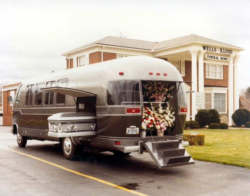 A converted RV with a rear flower display parked outside a funeral home, with a side door open, revealing its interior.