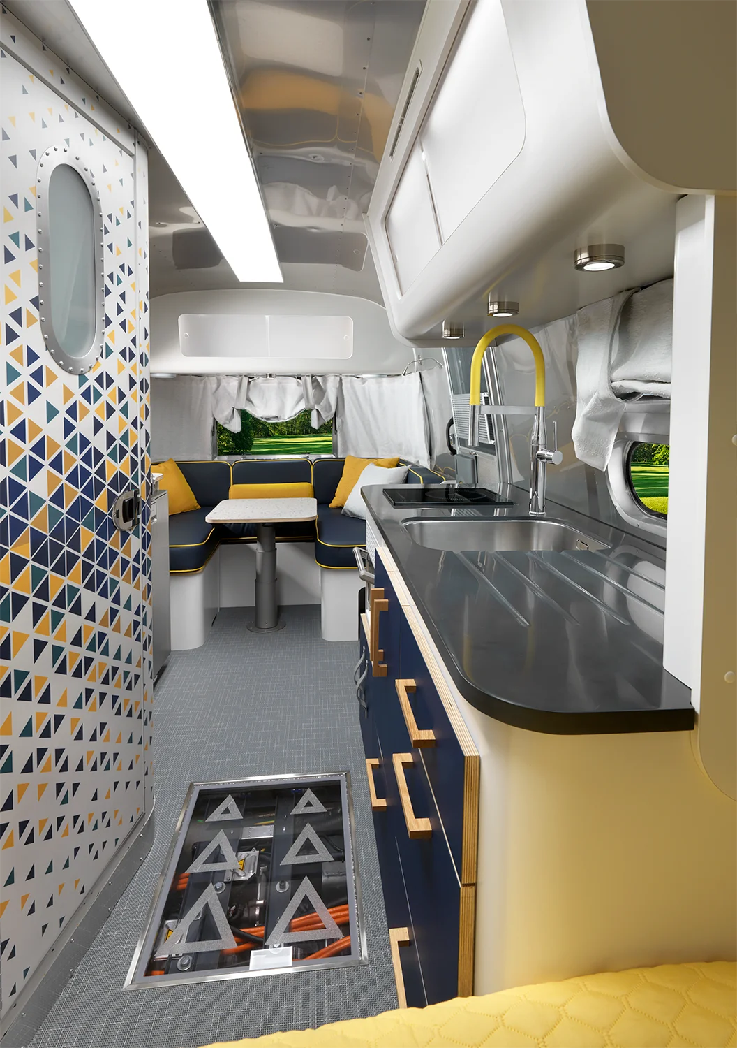 Modern RV interior with a sleek kitchen, gray countertops, yellow accents, a geometric-patterned door, a booth dining area with windows, and a glass floor panel revealing storage space below.