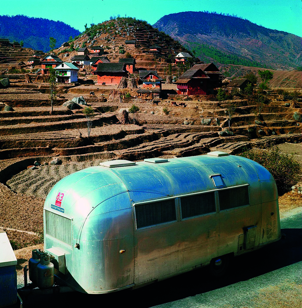 A shiny silver trailer is parked on a road next to terraced fields with scattered red houses and trees in a hilly, rural landscape under a clear blue sky.