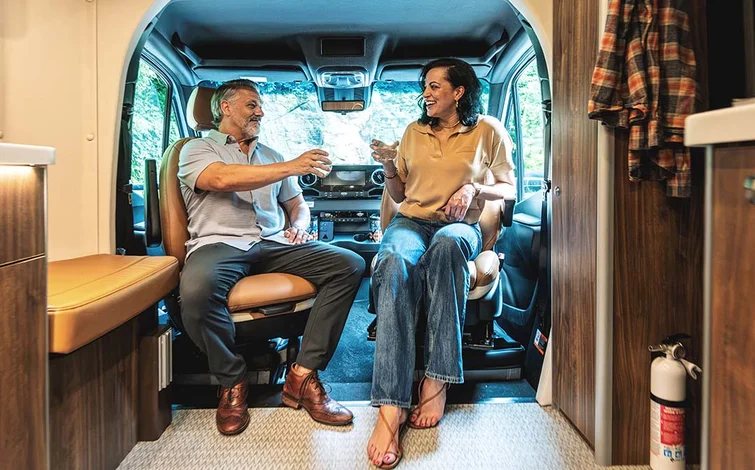 A man and woman sit facing each other in a camper van, holding drinks and smiling. They are surrounded by wood-paneled walls and soft lighting.