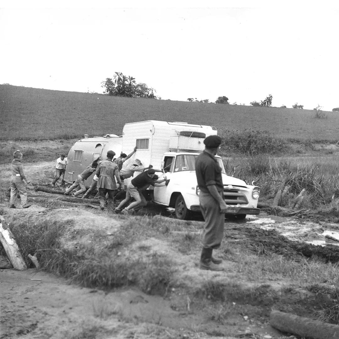 A group of people push a truck towing a caravan out of muddy terrain, while another person observes in the foreground.