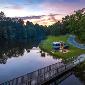 A camper van parked near a serene lake at sunset, with a picnic setup and paddleboards on the grass surrounded by lush trees.