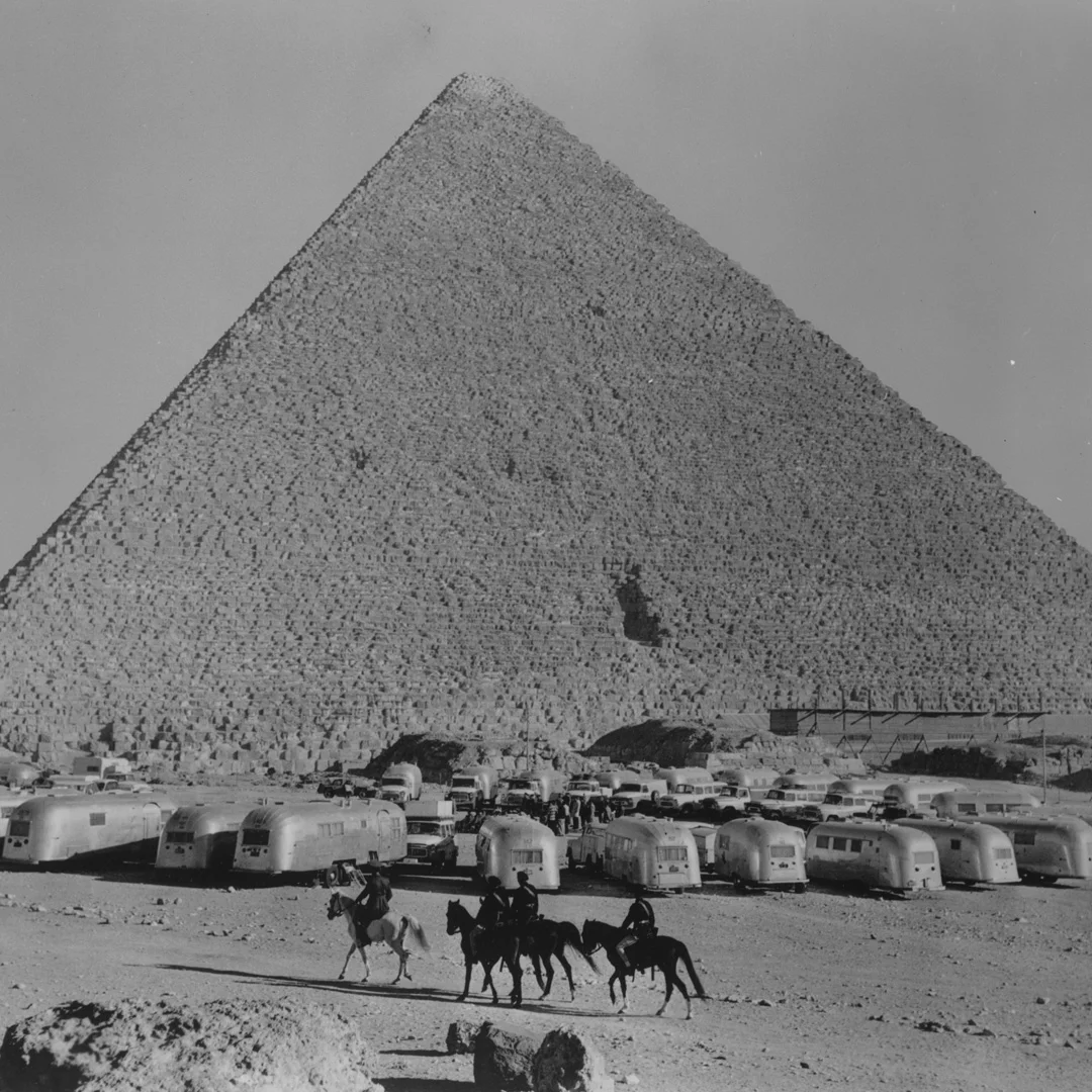 The Great Pyramid of Giza in the background with rows of Airstream trailers and three people on horseback in the foreground, black and white.