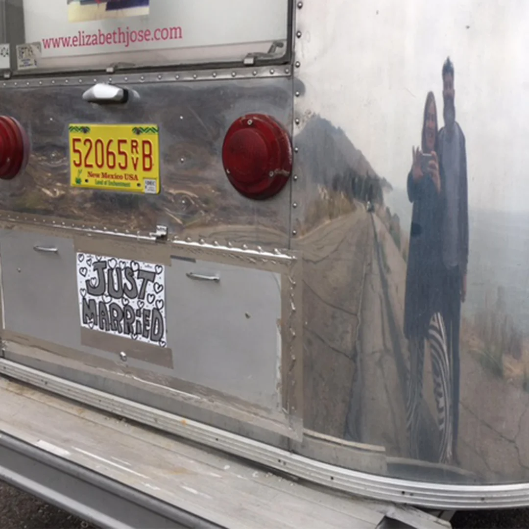 The back of a reflective trailer with "Just Married" sign and two people's reflection, parked by a road, showing mountains in the distance.