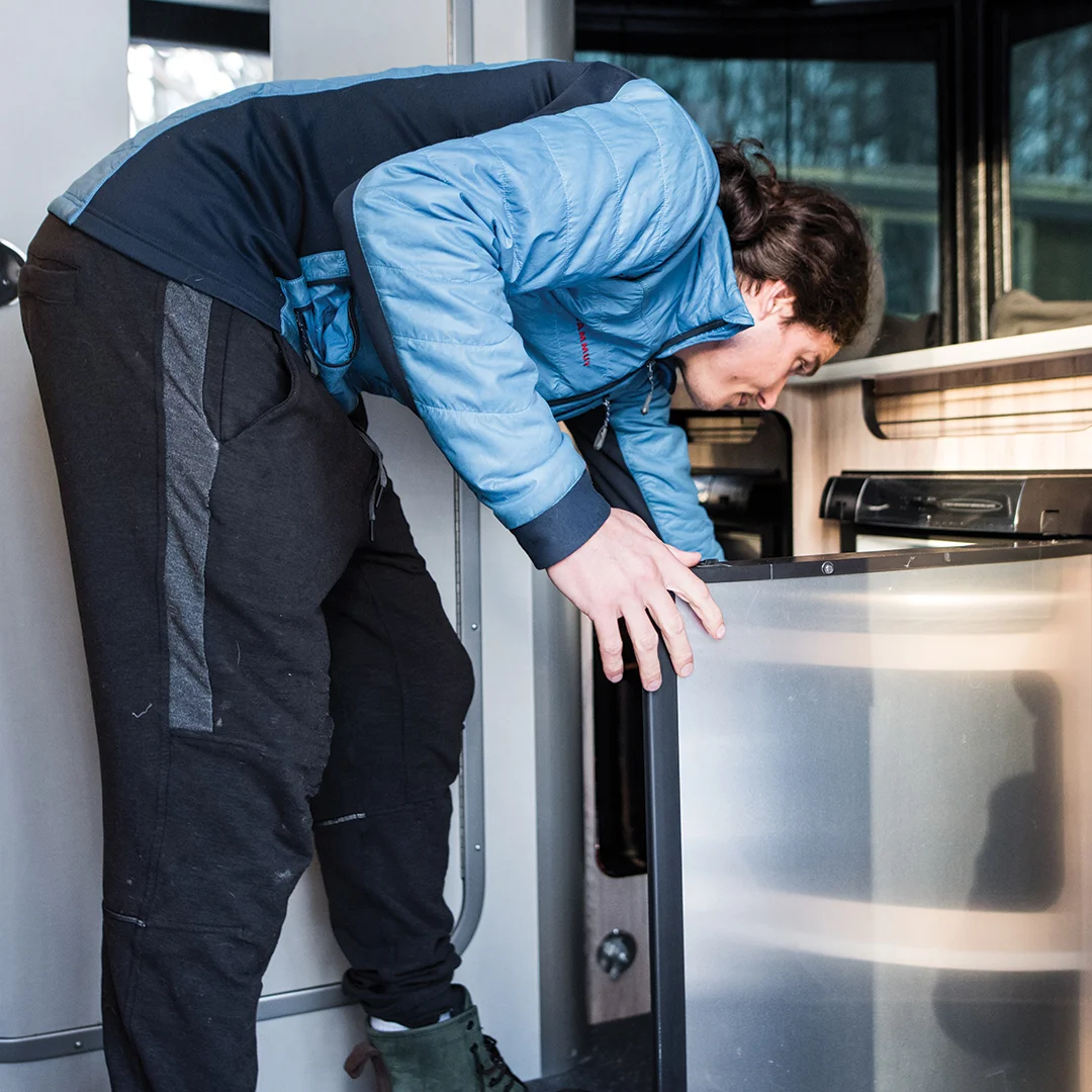 Person in a blue jacket and black pants checking a mini-fridge inside a camper van.