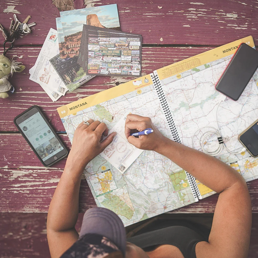 Person planning a trip using a Montana map, with postcards, a phone, and a tablet on a purple wooden table.