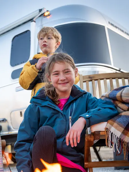Two children enjoy a campfire in front of an Airstream trailer. The girl smiles in a blue jacket, while the boy stands behind in a yellow coat.