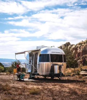 A silver Airstream trailer is parked in a scenic desert landscape with rocky formations. A person is seated outside at a small table.