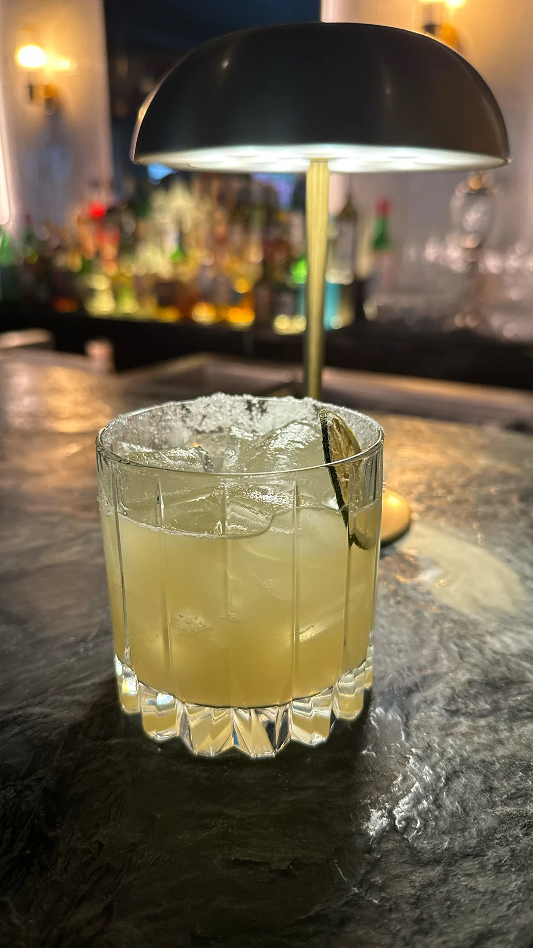 A frosty cocktail with a lime slice sits on a dimly lit bar counter, next to a sleek lamp with warm ambient light.