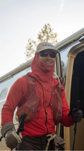 Person in hiking gear, wearing a white cap and sunglasses, smiles while holding trekking poles beside a trailer in a wooded area.