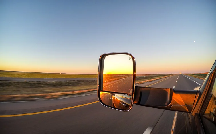 Sunset reflected in a car side mirror on an open highway, with vast fields and a clear sky in the background.