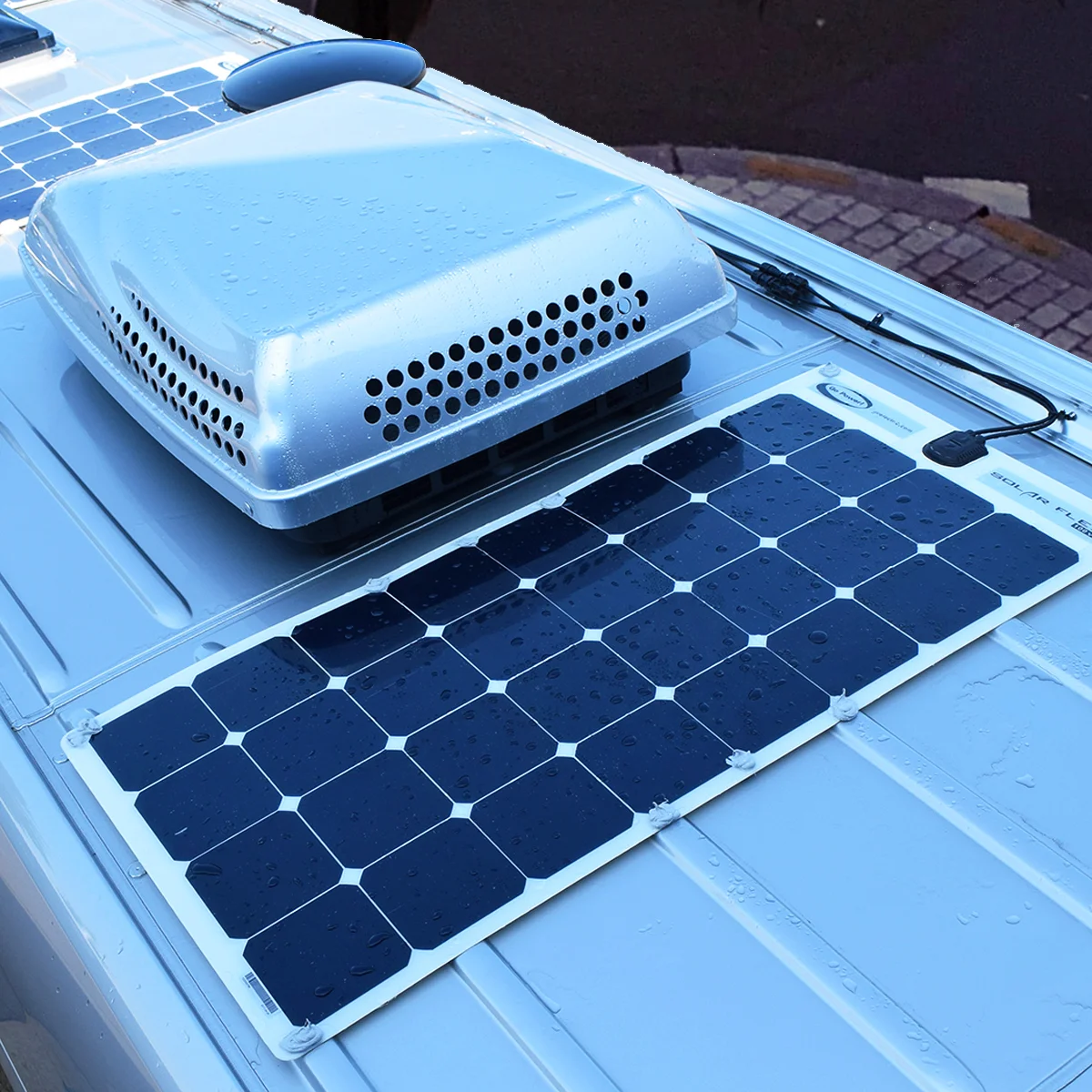 A flexible solar panel with water droplets is installed on the roof of a vehicle next to a rooftop ventilation unit. The panel is connected by a cable, and bricks are visible in the background.