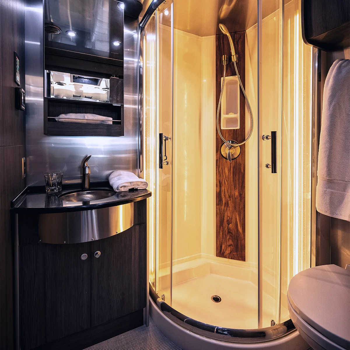 Modern bathroom with a curved glass shower, wood accents, overhead lighting, a black vanity with a sink, mirror cabinet, white towels, and a toilet in the foreground.
