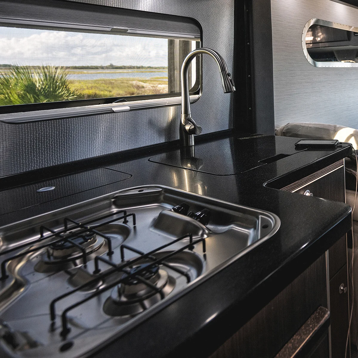 Compact camper van kitchen with black countertop, two-burner gas stove, and stainless steel faucet. Window reveals grassy landscape with water.