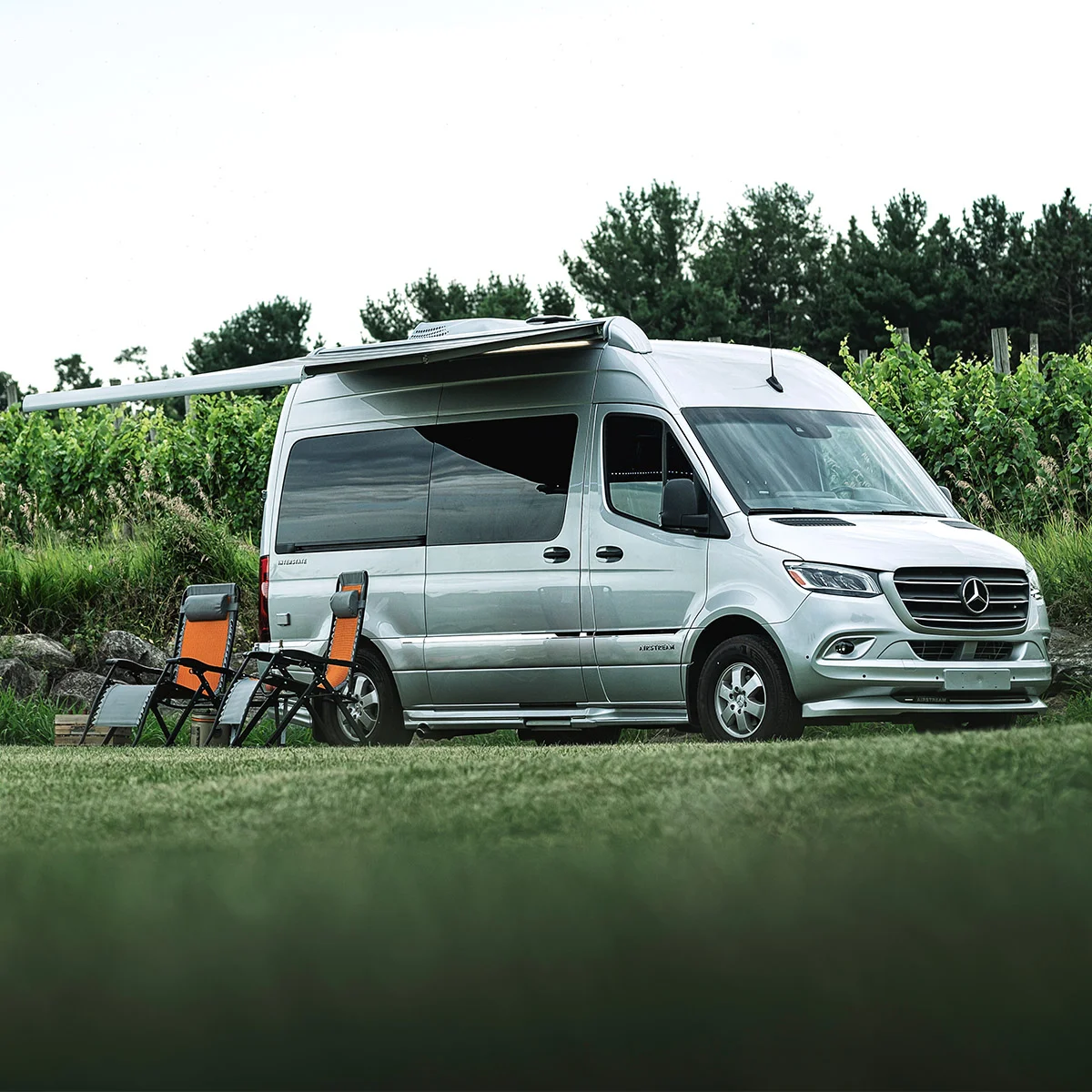 Silver camper van parked on grassy field with two orange and black chairs beside it, surrounded by lush greenery and trees.