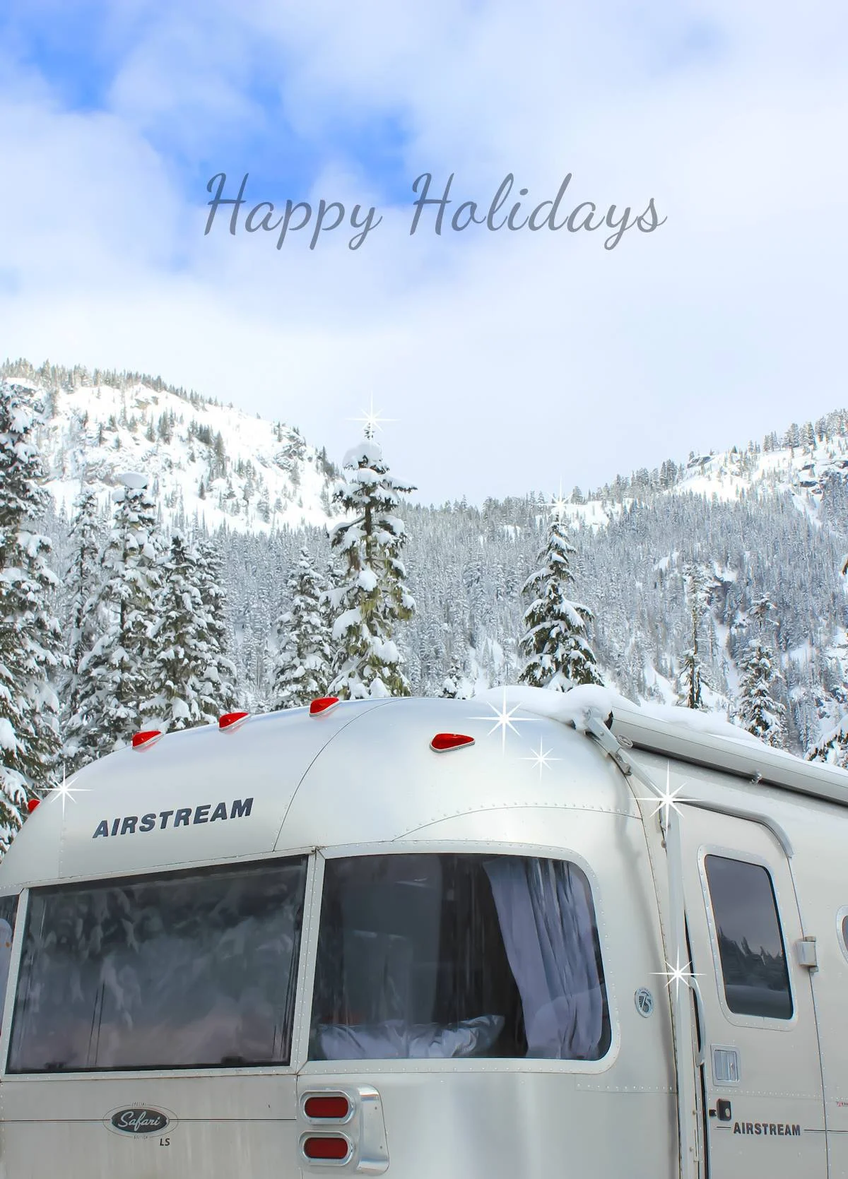Airstream trailer in snowy mountain landscape with "Happy Holidays" text above, surrounded by snow-covered trees and clear blue sky.