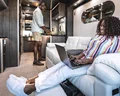 A woman works on a laptop while reclining on a sofa in a modern RV, as a man prepares food in the kitchen area.