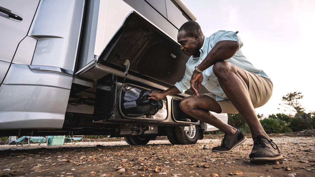 A man kneels beside an RV, placing a bag into a storage compartment. He is outdoors on a gravel surface, wearing a light shirt, shorts, and dress shoes. Trees and camping chairs are visible in the background.