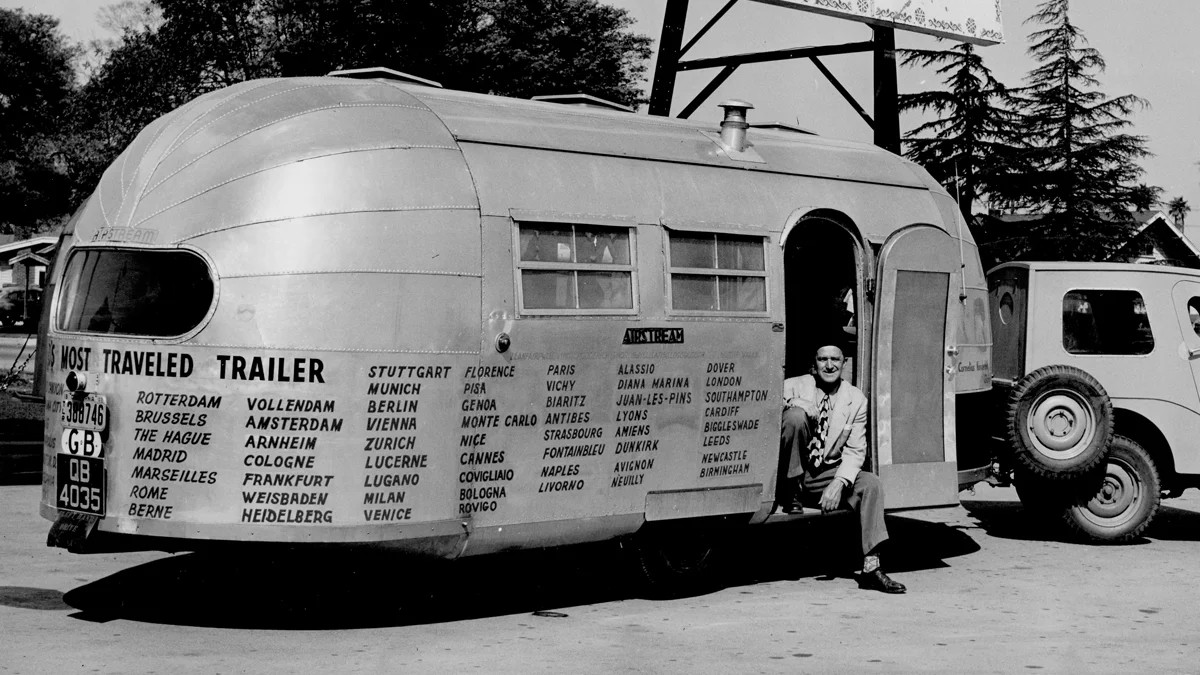 A vintage trailer with travel destinations listed is parked beside a jeep. A man sits in the trailer's doorway, smiling.