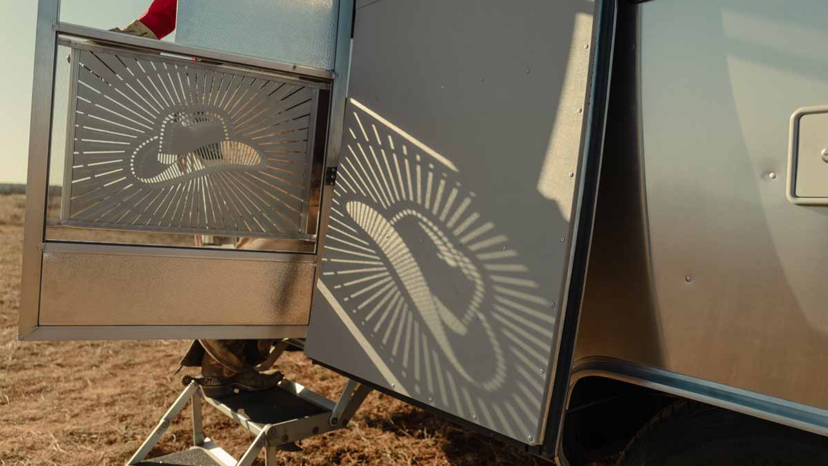 Shadow of a cowboy hat design cast on a trailer door, with a person in red behind an open screen door, steps leading down to brown grass.