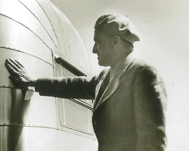 A man wearing a suit and beret stands in profile, touching the curved, metallic surface of an airplane or similar structure, with visible rivets and panels, in a black-and-white photograph.