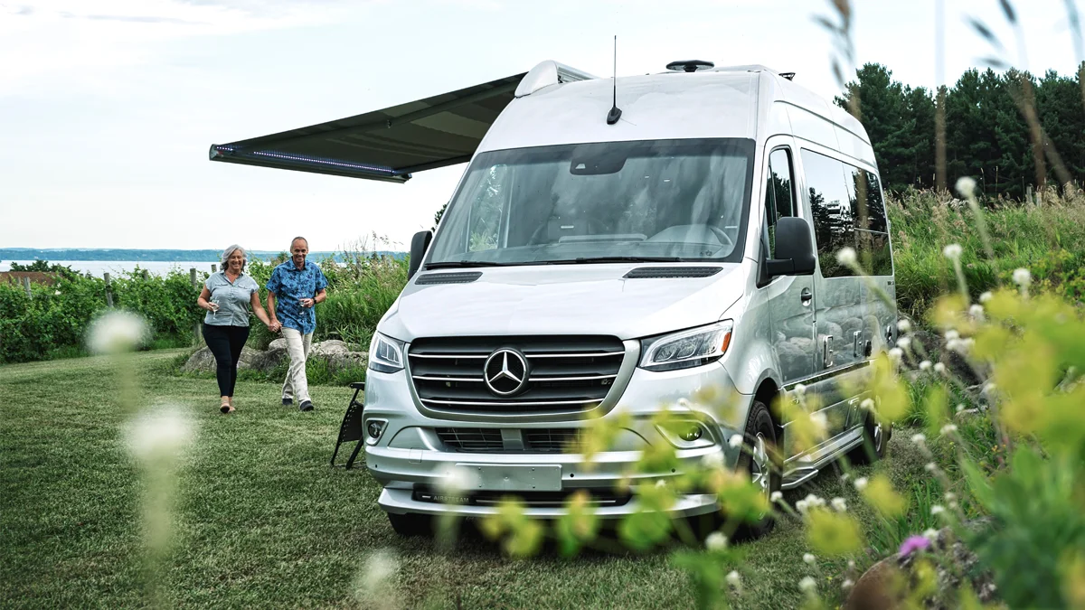 A couple walks beside a parked Mercedes camper van with an awning extended, set in a grassy area with trees and a lake in the background.