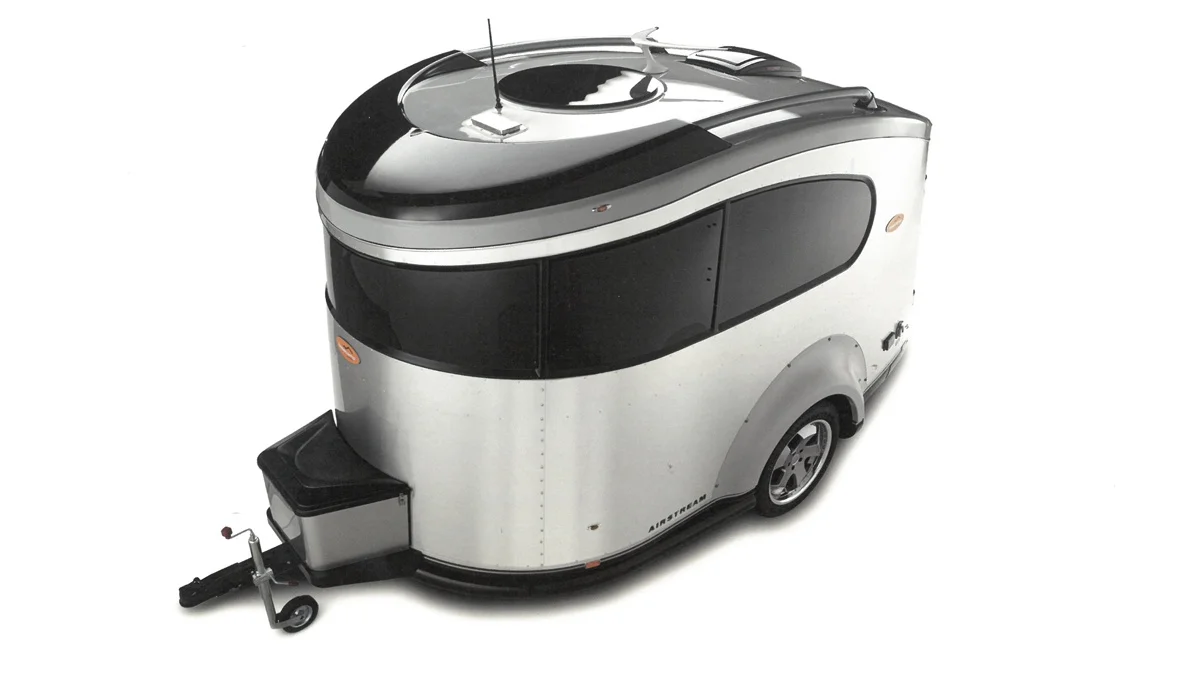 A modern, compact silver travel trailer with black accents, rounded edges, a single axle, and tinted windows, shown isolated on a white background.