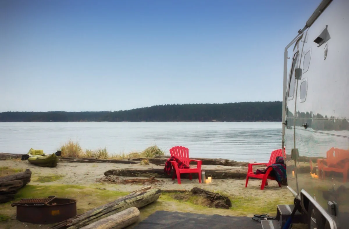 Camping scene by a calm lake with red chairs, a kayak, and an RV. Tranquil setting with logs and grass, under a clear blue sky.
