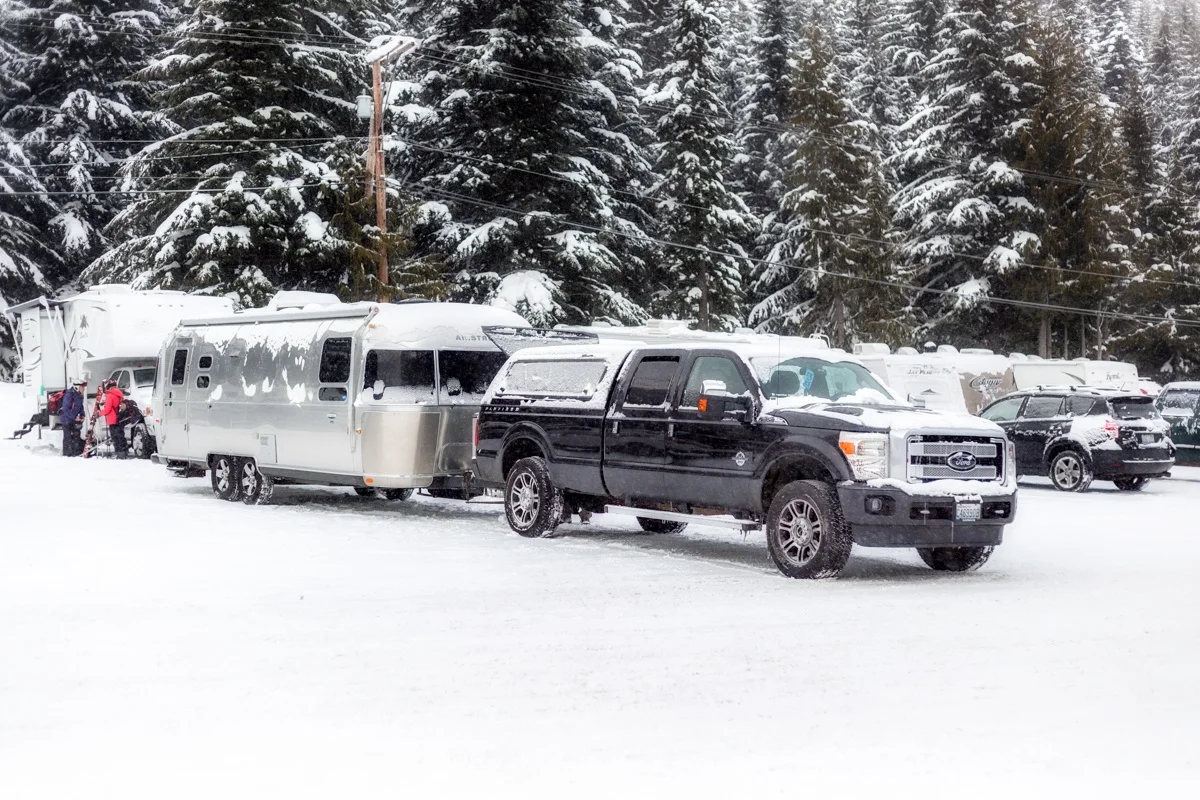 A black truck with a silver trailer in a snowy parking area, surrounded by trees covered in snow.