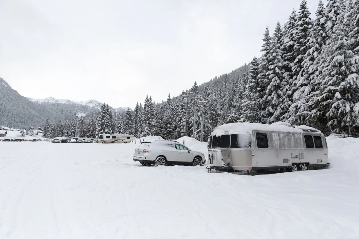 Snow-covered camper and cars in a wintery mountain landscape with dense, snow-laden evergreen trees and a cloudy sky.