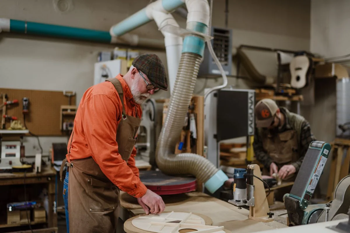 Two people work in a woodshop; the man in front, wearing an orange shirt, apron, and cap, shapes wood at a workbench, while another person works on machinery in the background. Various woodworking tools are visible.
