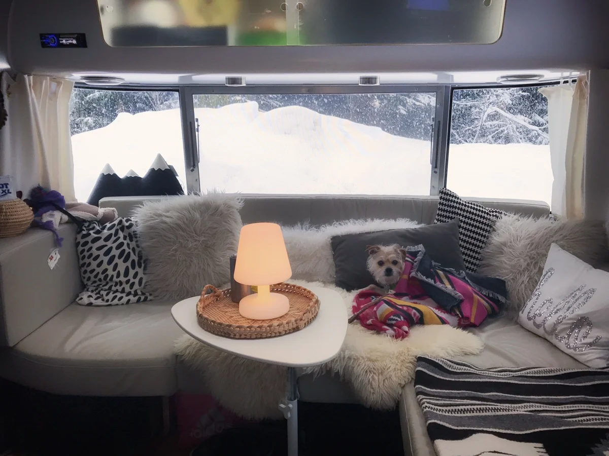 Cozy camper interior with a small dog wrapped in a colorful blanket on a sofa. A glowing lamp sits on a table, and snow is visible through the window.