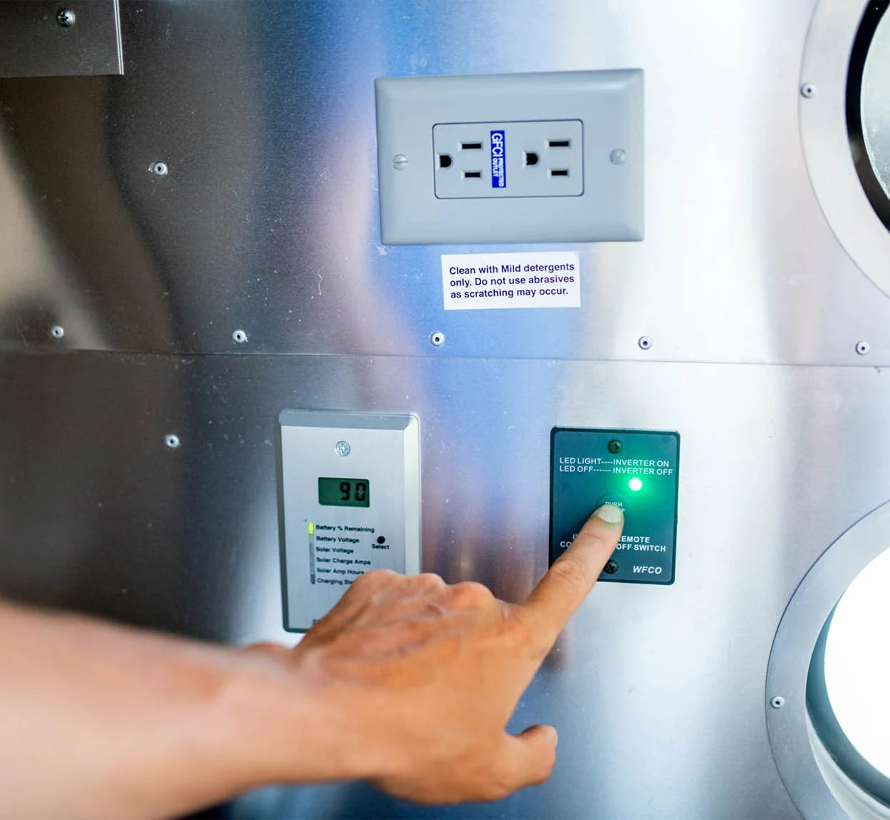 A hand presses a green-lit button on a metal panel beneath a digital display and electrical outlet, with an instruction label above the outlet.