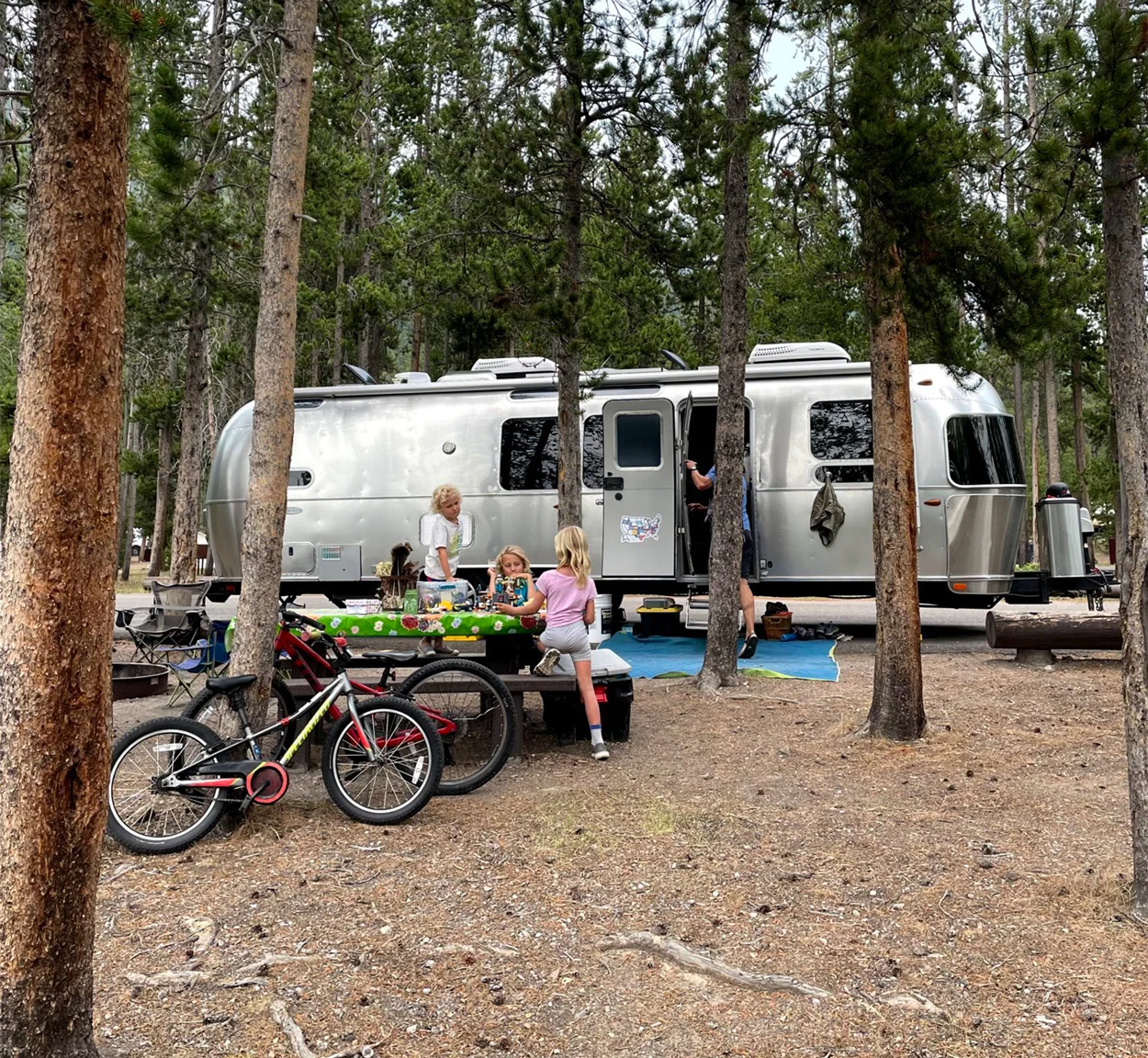Three kids play at a picnic table in front of a silver camper trailer parked among tall trees. A bicycle leans nearby, and an adult stands at the trailer door. The scene appears to be a family camping trip.