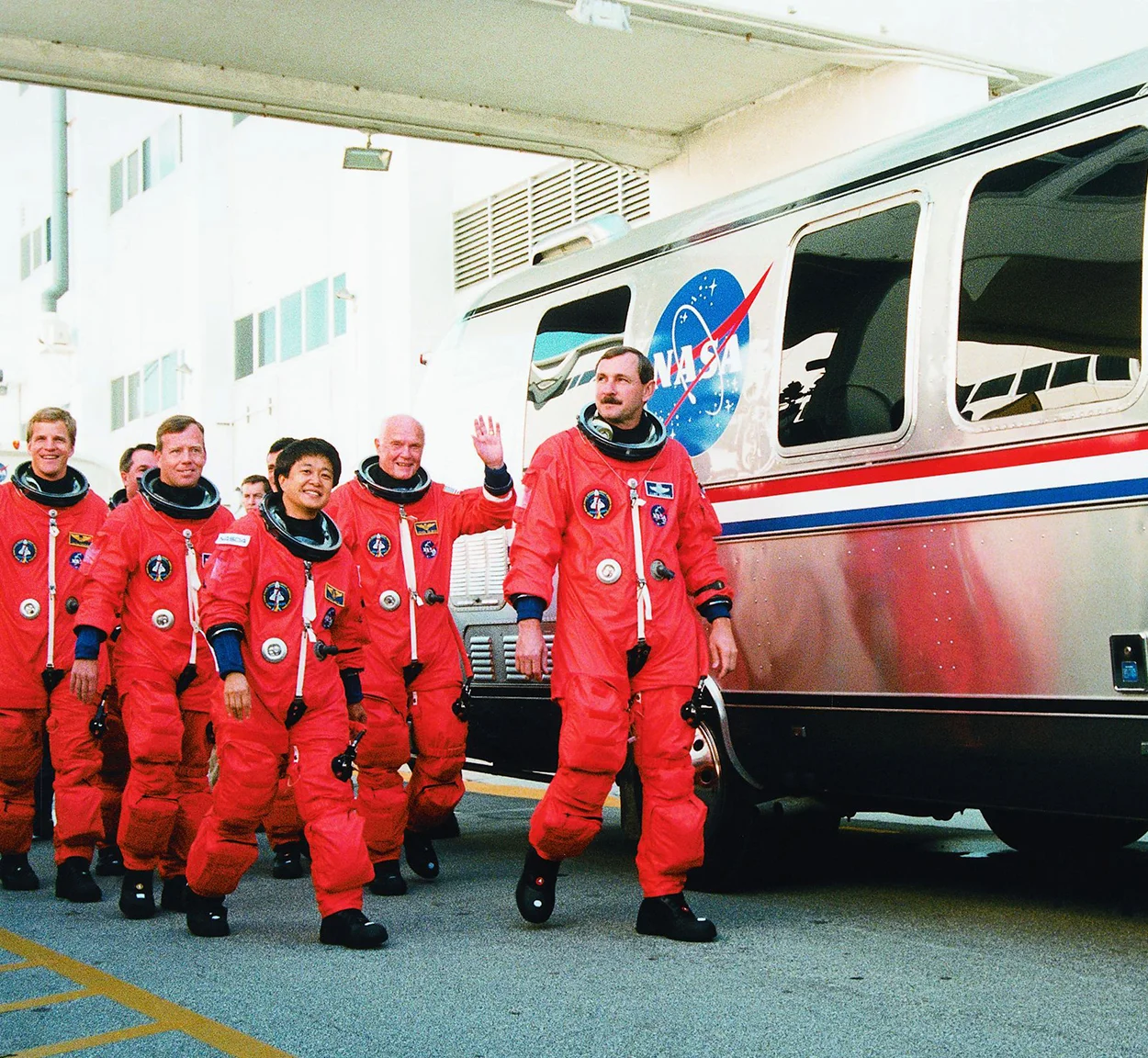 A group of astronauts in orange space suits walks beside a NASA vehicle, smiling and waving, while preparing for a space mission.