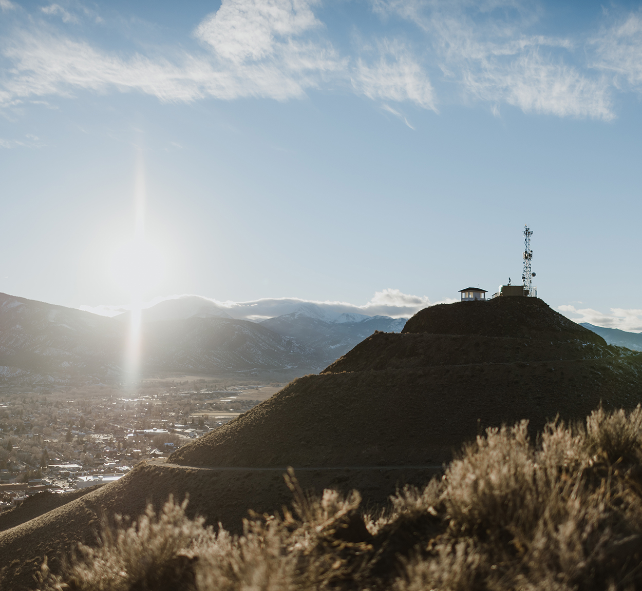 The sun rises over distant mountains and a small valley town, with a radio or cell tower and a small building on top of a hill in the foreground. Sparse vegetation covers the hills under a partly cloudy sky.