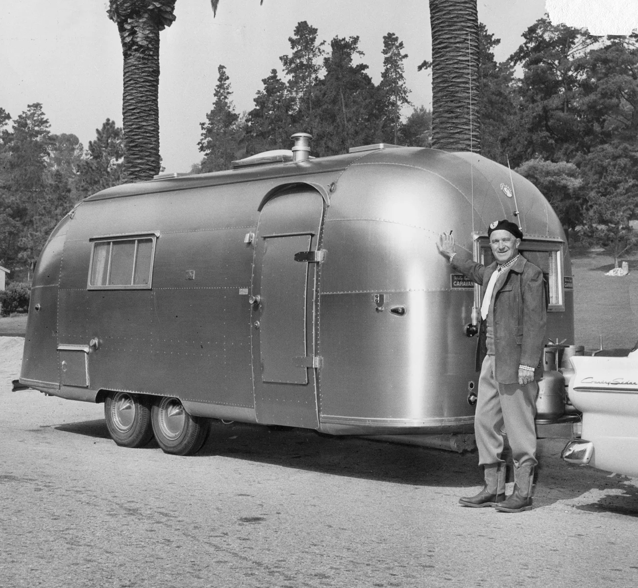 Man standing beside a vintage aluminum travel trailer hitched to a car, with palm trees and forested area in the background.