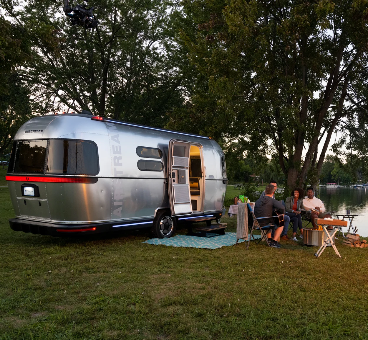 A silver Airstream trailer is parked on grass by a lake. Four people sit around a table and campfire, talking and enjoying the outdoors, surrounded by trees and green grass.