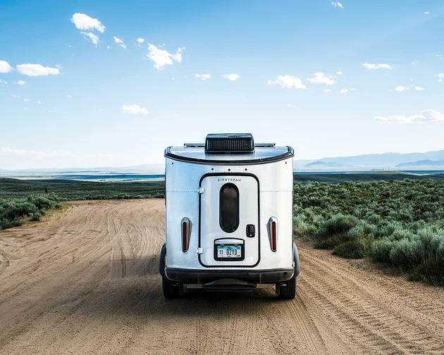 A silver travel trailer drives down a dirt road surrounded by green fields and distant mountains under a bright blue sky with scattered clouds.