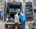 Two workers examine the interior wiring of a van inside a factory, with the back doors open and various cables exposed.