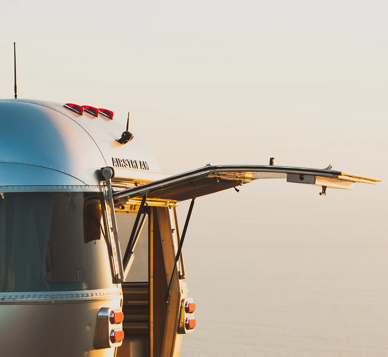 A close-up of an Airstream trailer with its rear hatch open, overlooking a calm, hazy ocean at sunset. The metallic surface reflects the warm, golden light.
