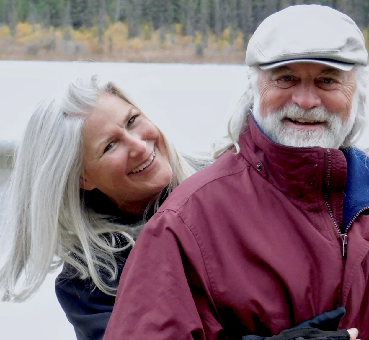 A smiling older couple with gray hair poses outdoors. The woman leans affectionately on the mans shoulder, and he wears a white flat cap and maroon jacket. Trees and a lake appear blurred in the background.