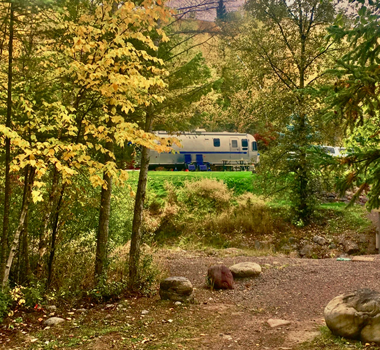 A silver camper trailer is parked on grass in a wooded area with yellow-green autumn leaves, two blue chairs nearby, and large rocks in the foreground.