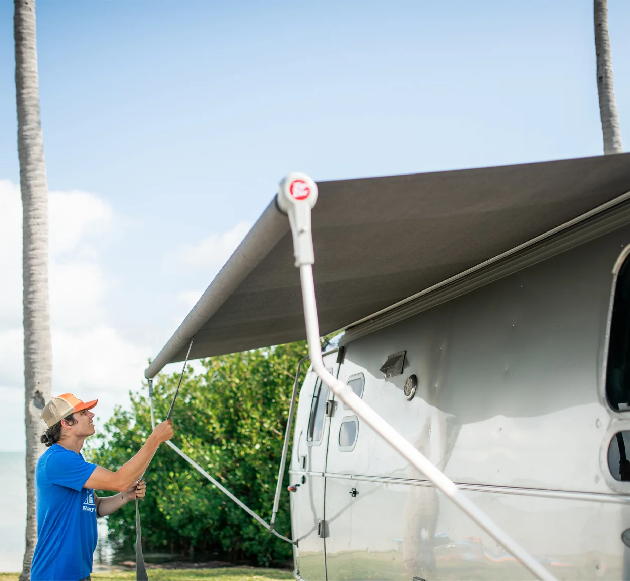 A person in a blue shirt and cap sets up a large gray awning attached to the side of a silver RV, with palm trees and greenery in the background.