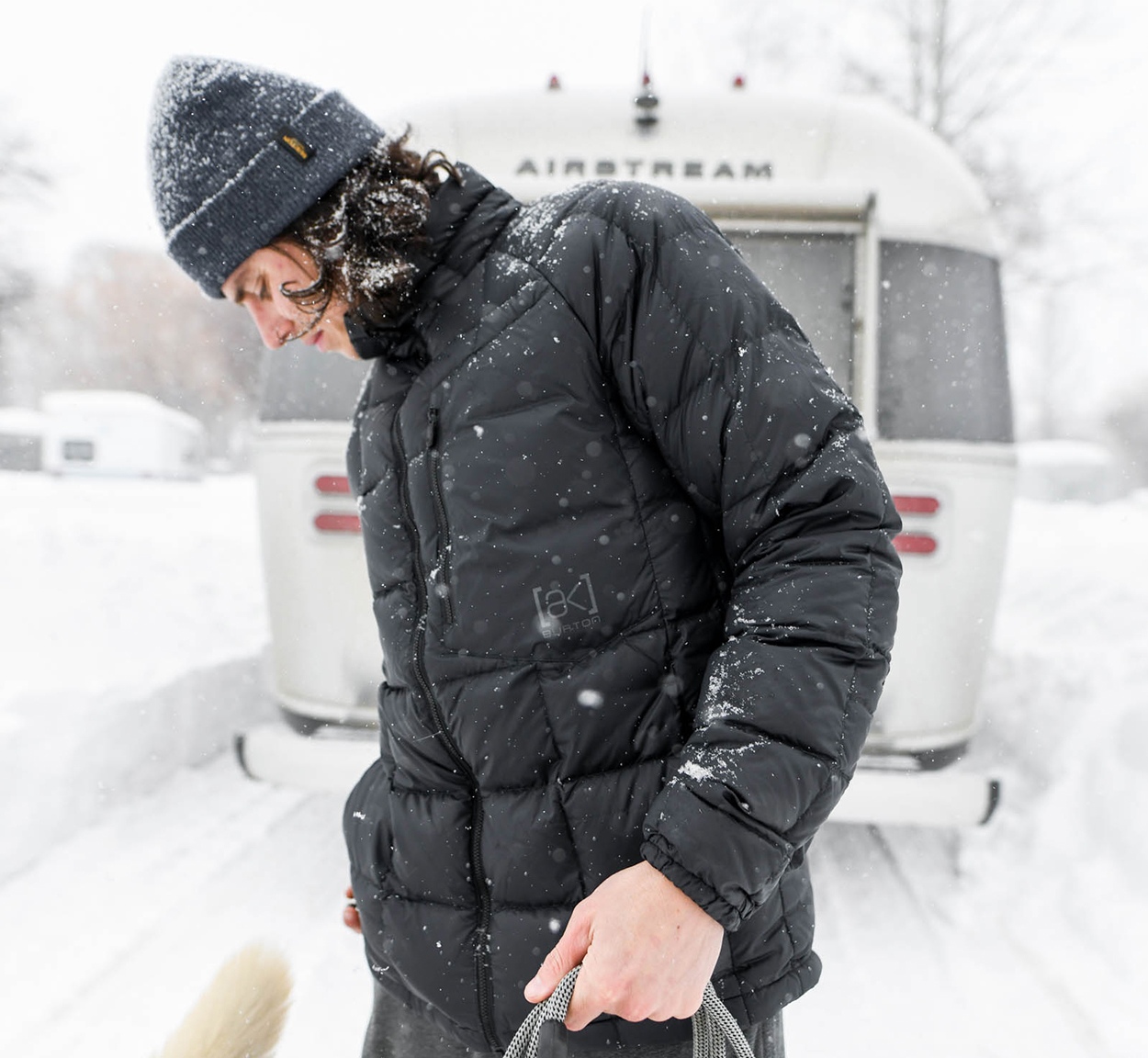 Person in a black winter jacket and gray beanie stands in a snowy scene in front of an Airstream trailer, holding a rope or leash.