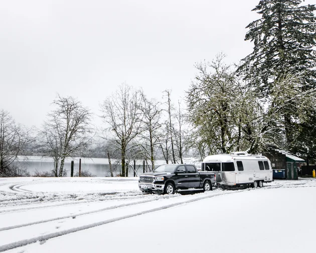 A pickup truck towing a silver travel trailer is parked on a snow-covered ground near a lake, with bare trees and tire tracks visible in the white landscape.