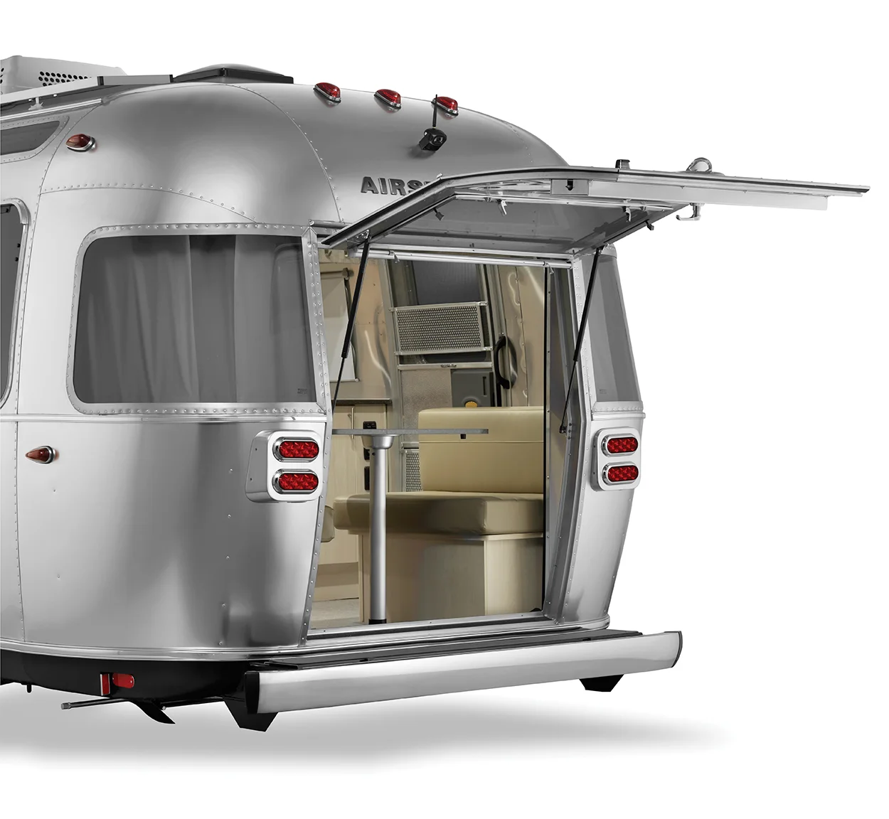 A silver Airstream trailer with its rear hatch open, revealing a modern, light-colored interior with seating and cabinetry. The trailer features rounded edges and classic vintage design elements.