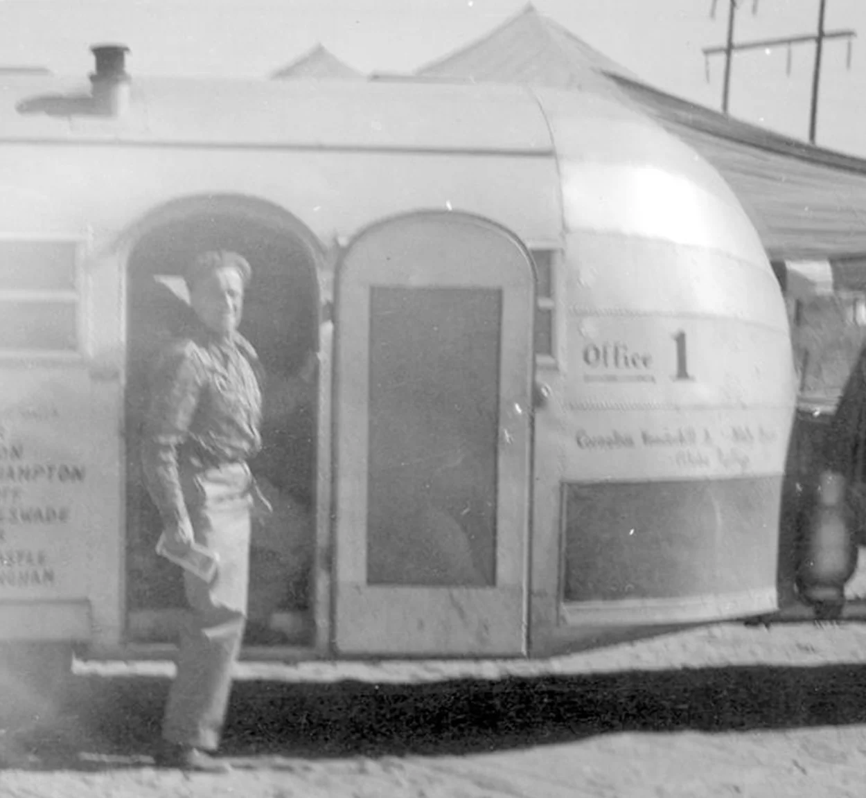 A person stands outside the door of a vintage trailer labeled Office 1, holding a piece of paper. The trailer is set up outdoors on sandy ground, with tents and utility poles in the background.