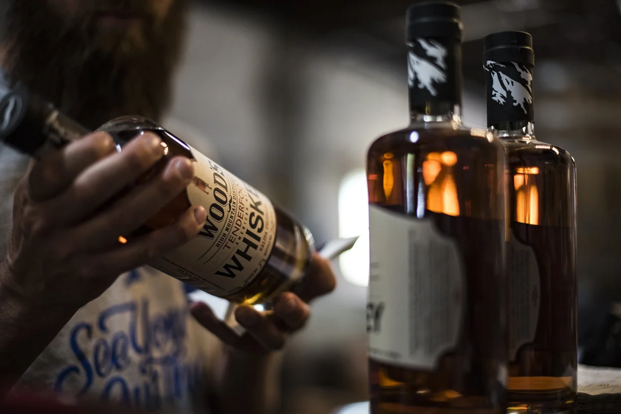 A person holds and examines a bottle of whiskey labeled Woodinville Whiskey Co., while two more whiskey bottles stand nearby on a table. The background is softly blurred.
